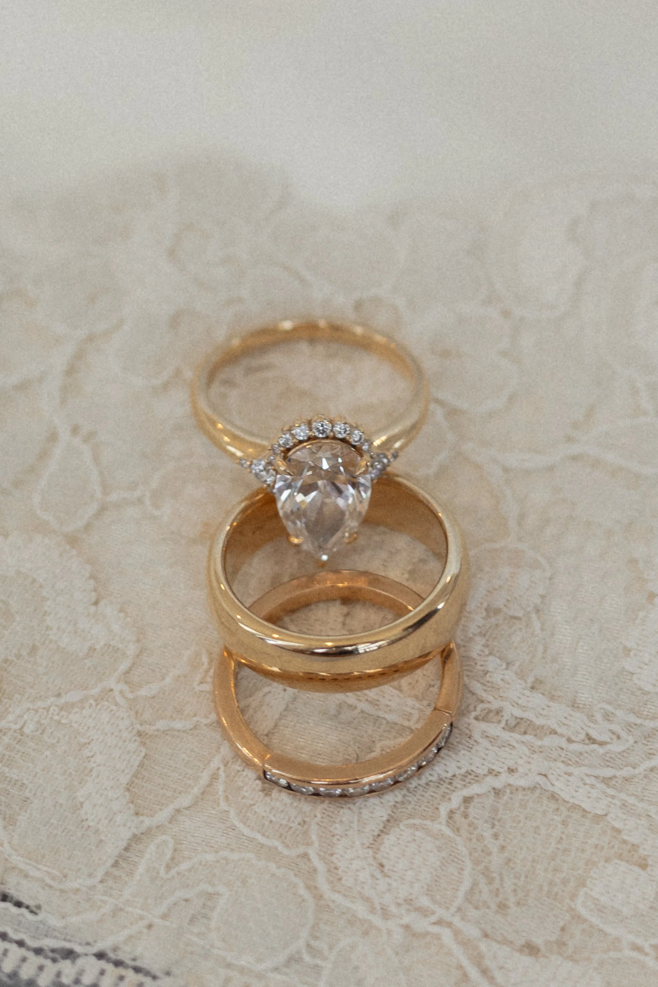 Stacked gold rings and a solitaire engagement ring with a large teardrop-shaped diamond on a lace tablecloth.