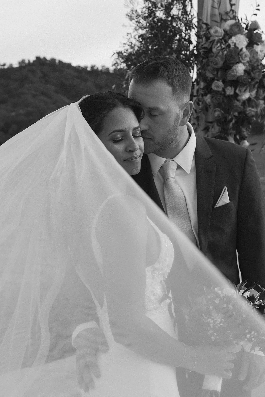 Black and white photo of a bride and groom close together, with the groom kissing the bride's temple. The bride is wearing a wedding dress and veil, holding a bouquet, and the groom is in a suit with a tie. There are flowers and open landscape in the background.