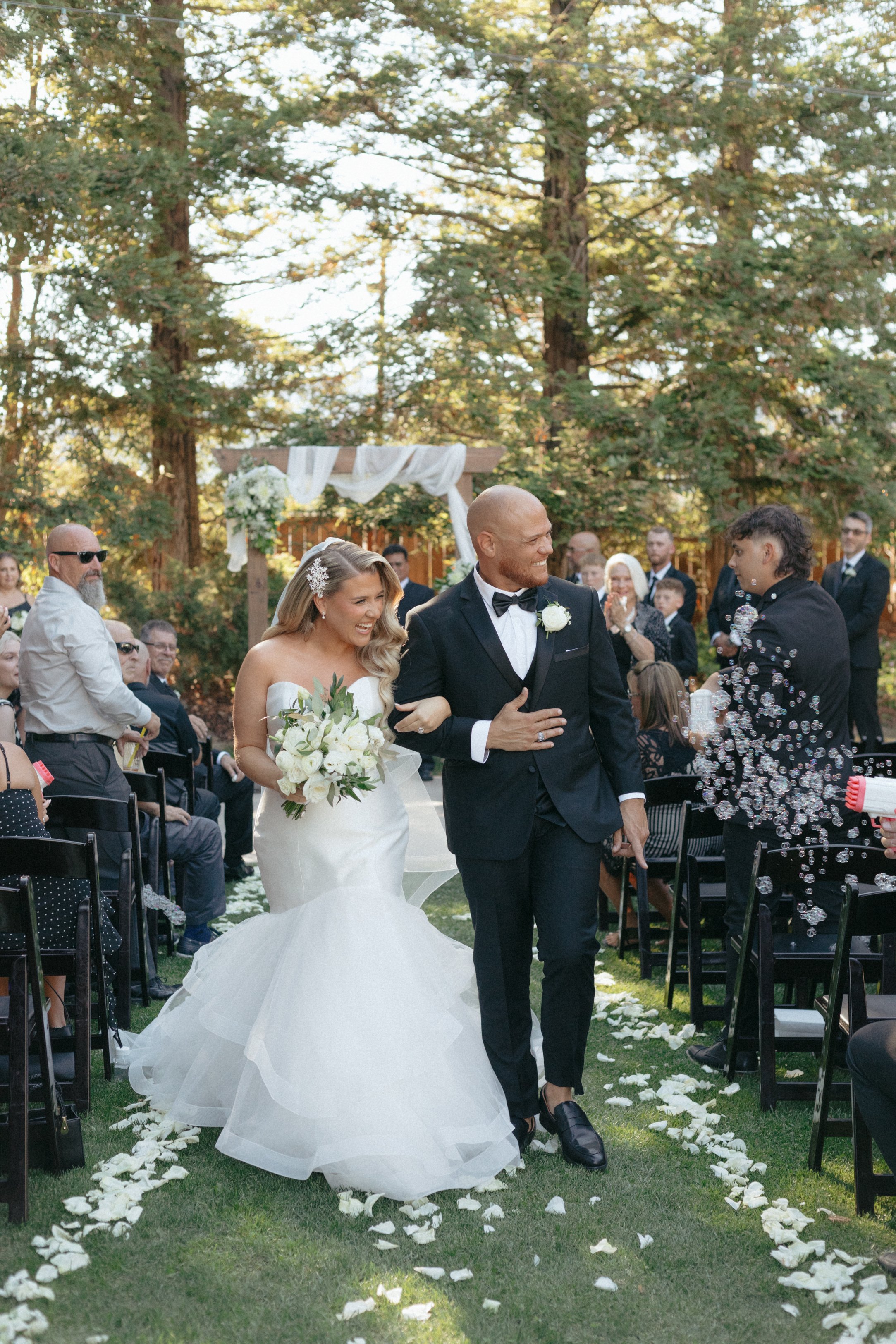 Couple walking down the aisle at an outdoor wedding ceremony, surrounded by seated guests, with a floral arch in the background, with pine trees and natural setting.