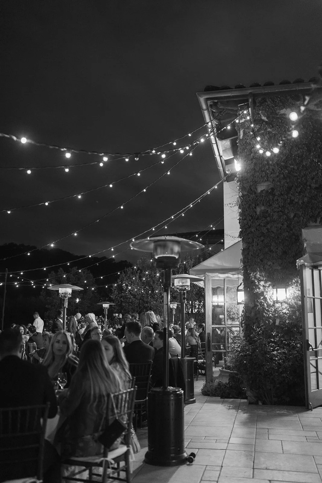 Night scene at an outdoor restaurant with tables, people dining, string lights overhead, and heaters providing warmth.