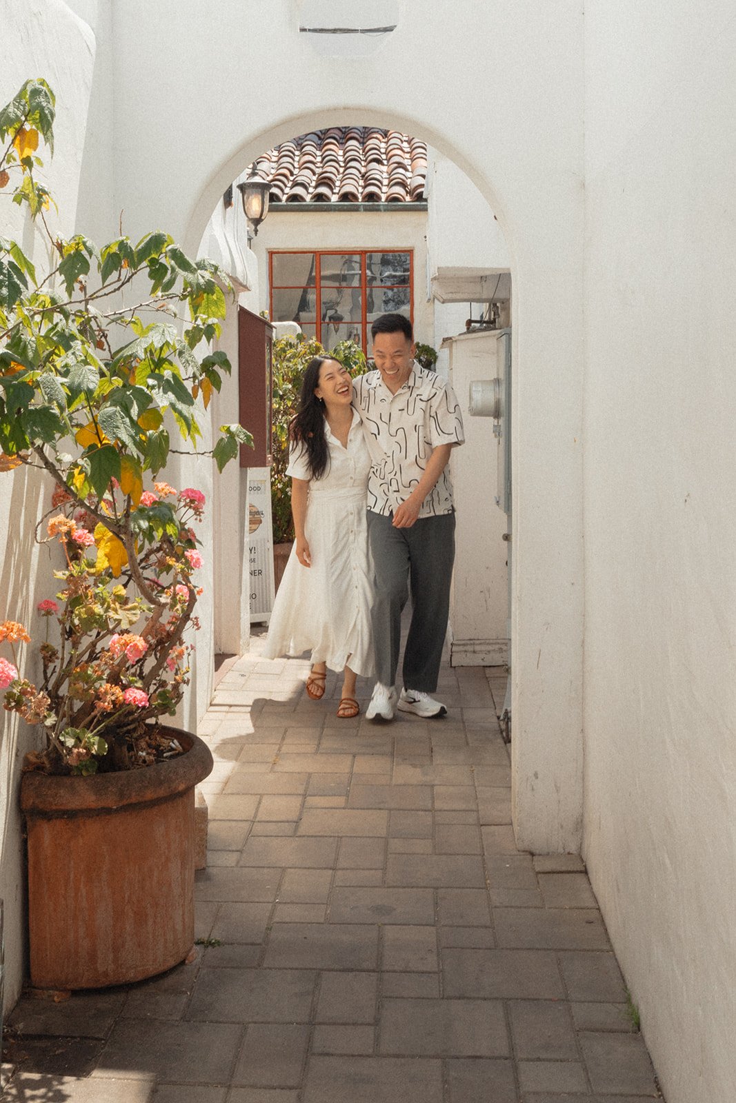 A young couple walking through a white archway, smiling and laughing, with a potted plant with pink and yellow flowers on the left, and a man and woman dressed in white and casual clothes, in a sunny outdoor setting.