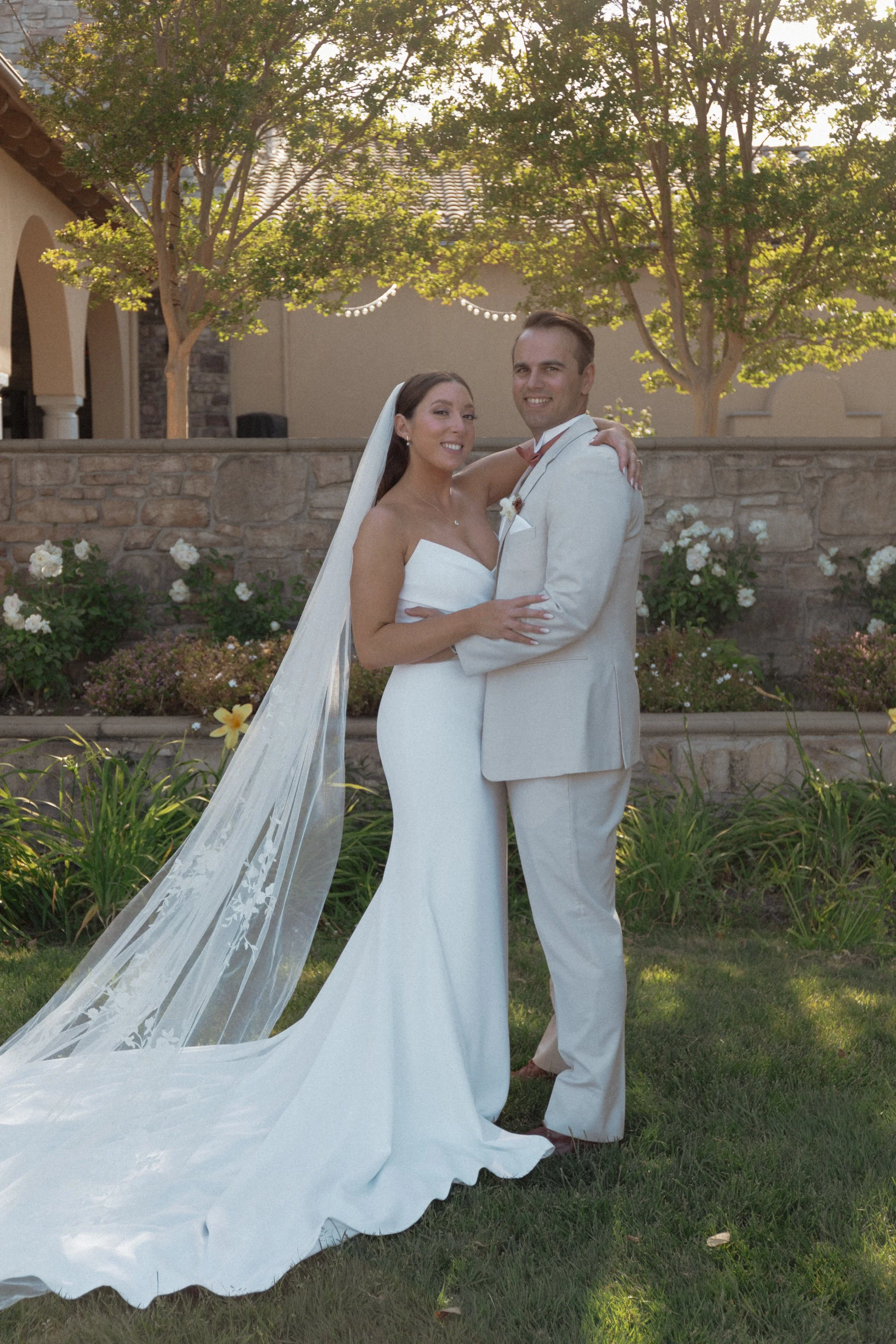 A bride and groom stand close together outdoors, embracing, during their wedding. The bride wears a white strapless wedding gown with a long lace veil, and the groom wears a light-colored suit. They are smiling, with a stone wall, greenery, and trees in the background.