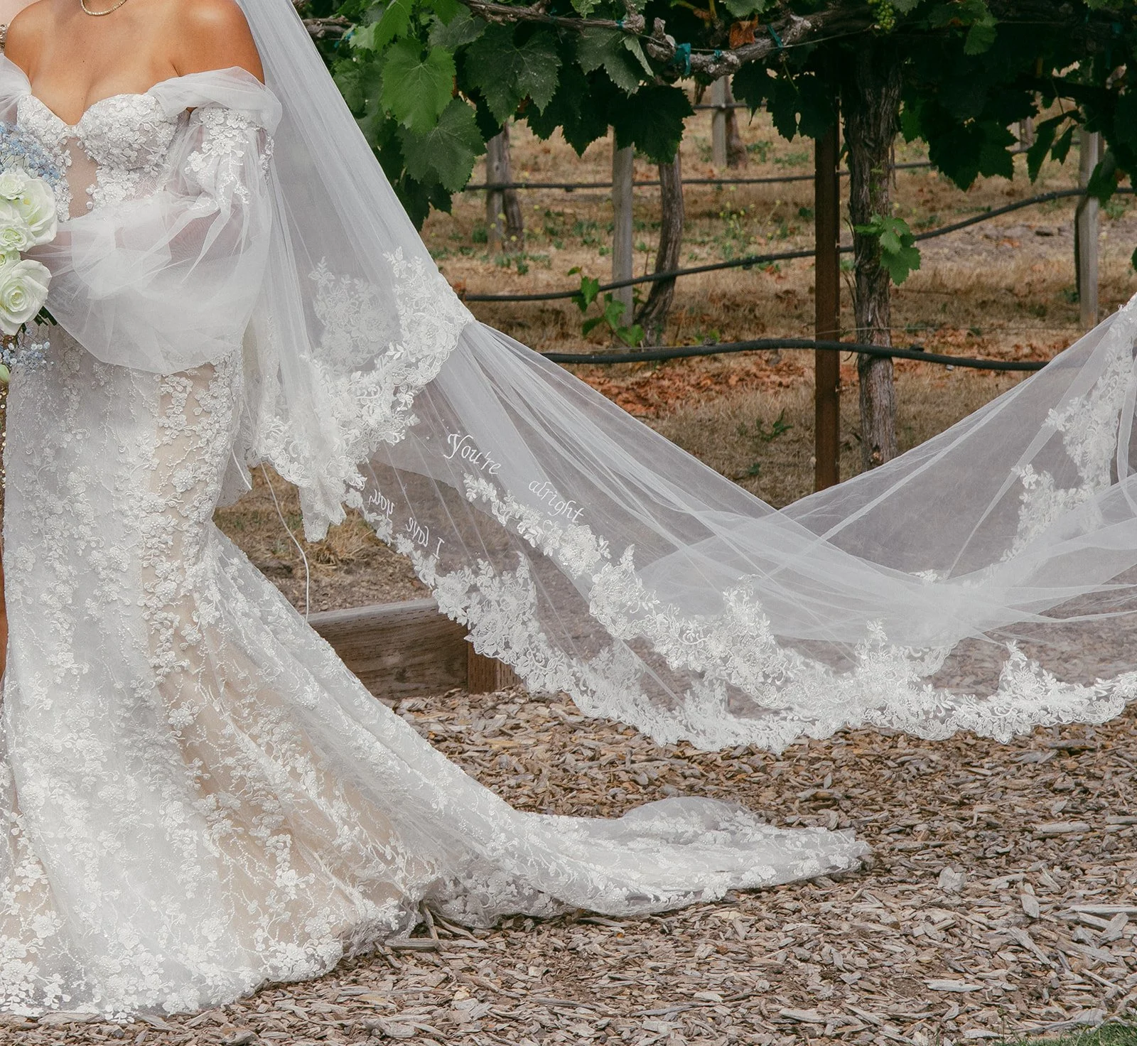 Close-up of a bride in a white lace wedding gown with a lengthy veil in a vineyard setting.