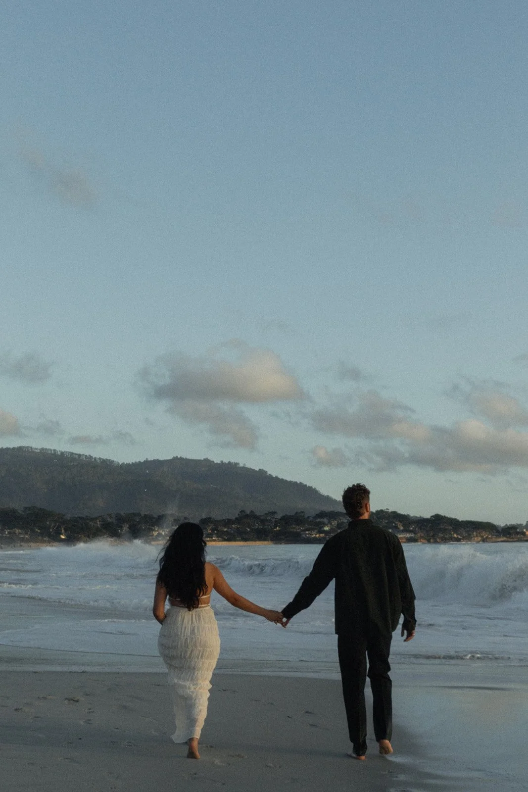 A couple holding hands while walking on the beach near the shoreline during dusk or early evening, with mountains and clouds in the background.