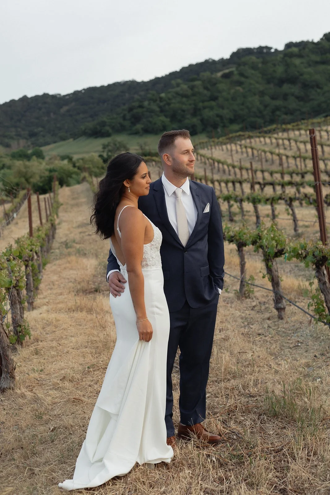Newlywed couple standing in a vineyard, with rolling hills and vines in the background during daytime.