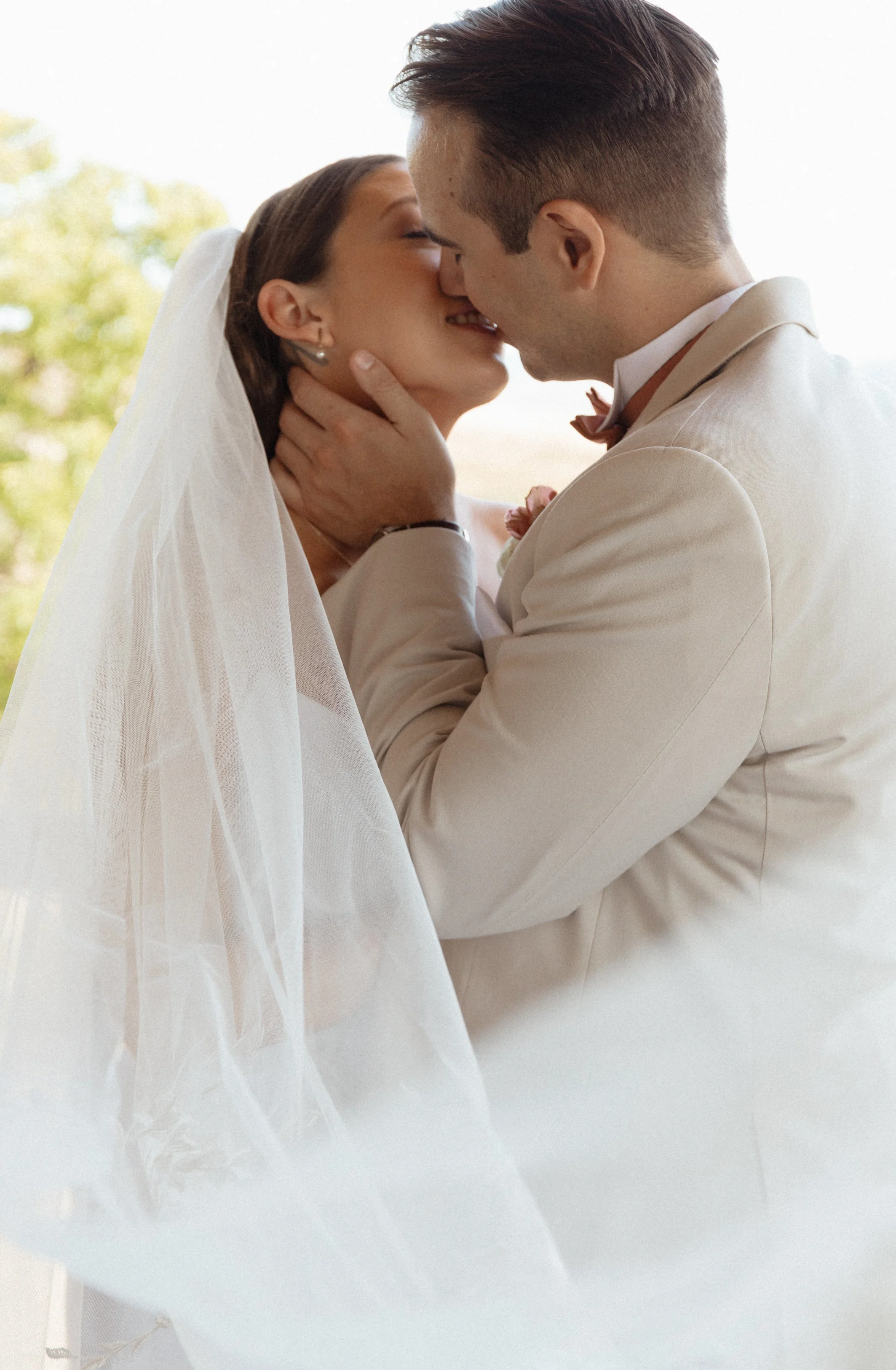 A bride and groom kissing outdoors, with trees in the background, the bride is wearing a veil and the groom is in a beige suit.