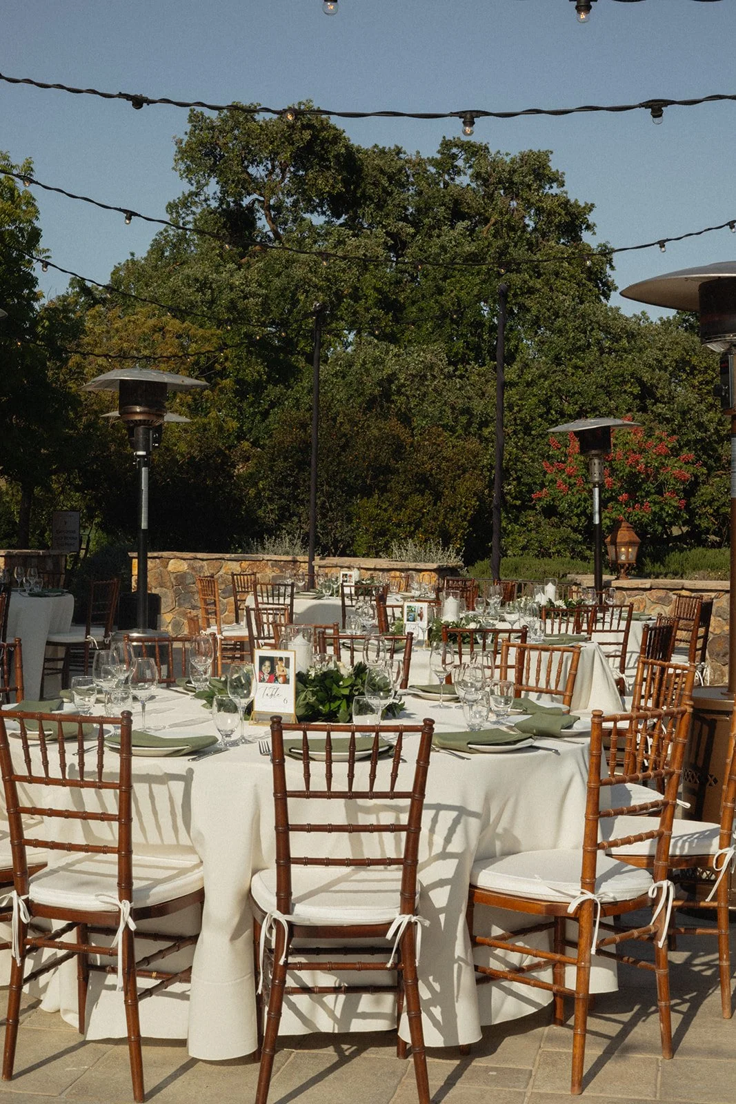 Outdoor wedding reception setup with round tables, white tablecloths, glassware, and wooden Chiavari chairs, in a garden setting with trees and string lights overhead.