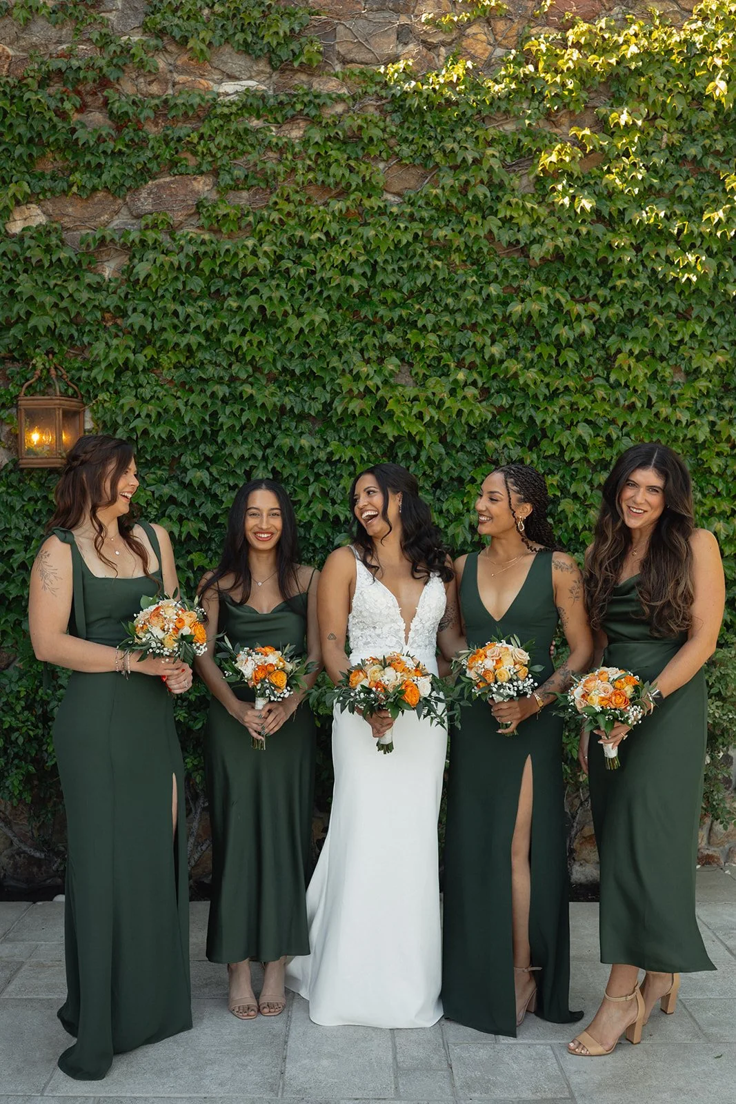 A bride in a white wedding dress stands with her four bridesmaids, all wearing matching dark green dresses, holding bouquets of orange and white roses. They are standing outdoors in front of a stone wall covered with green ivy, smiling and laughing together.