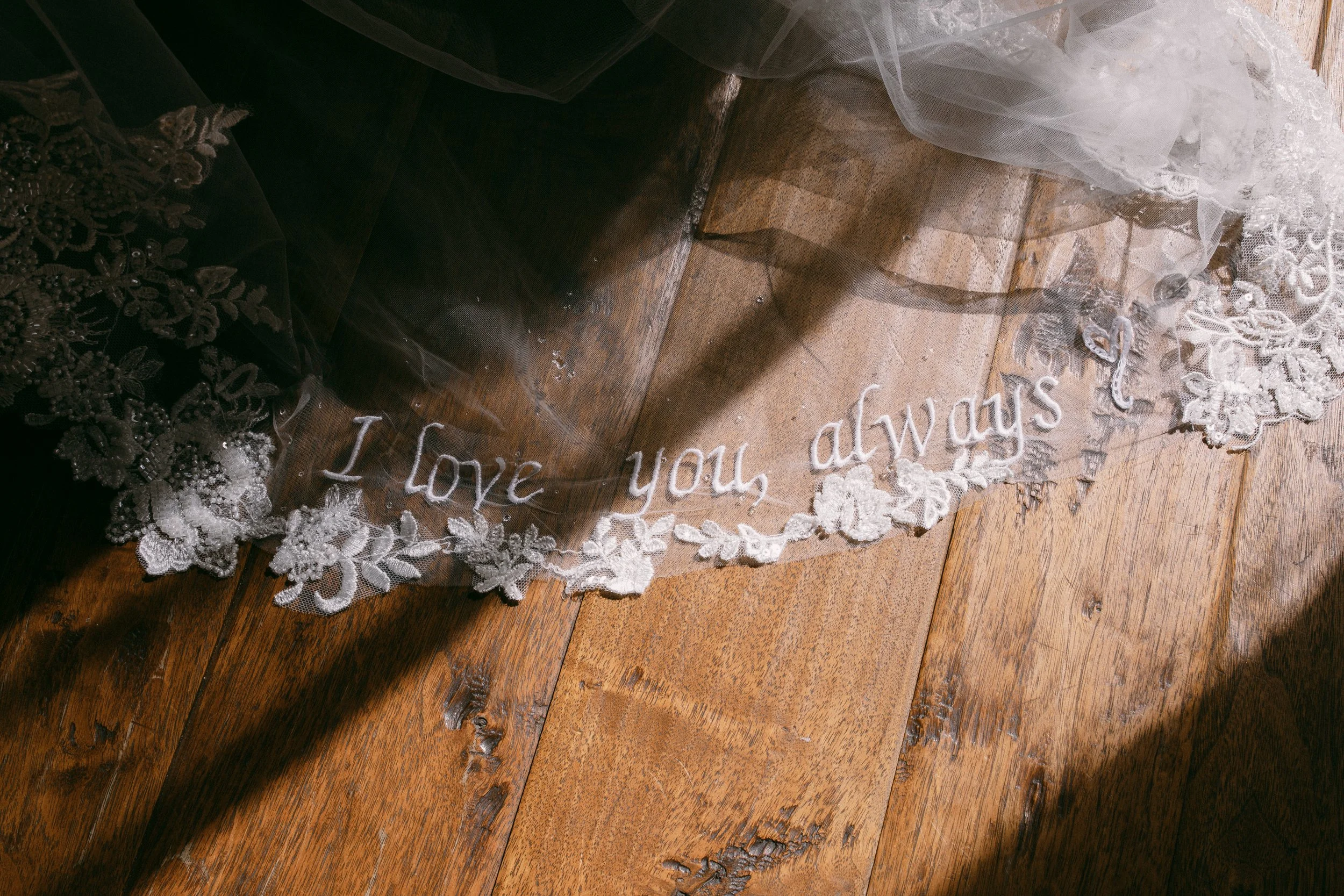 Close-up of a lace veil with the embroidered message "I Love you always" lying on a wooden floor.
