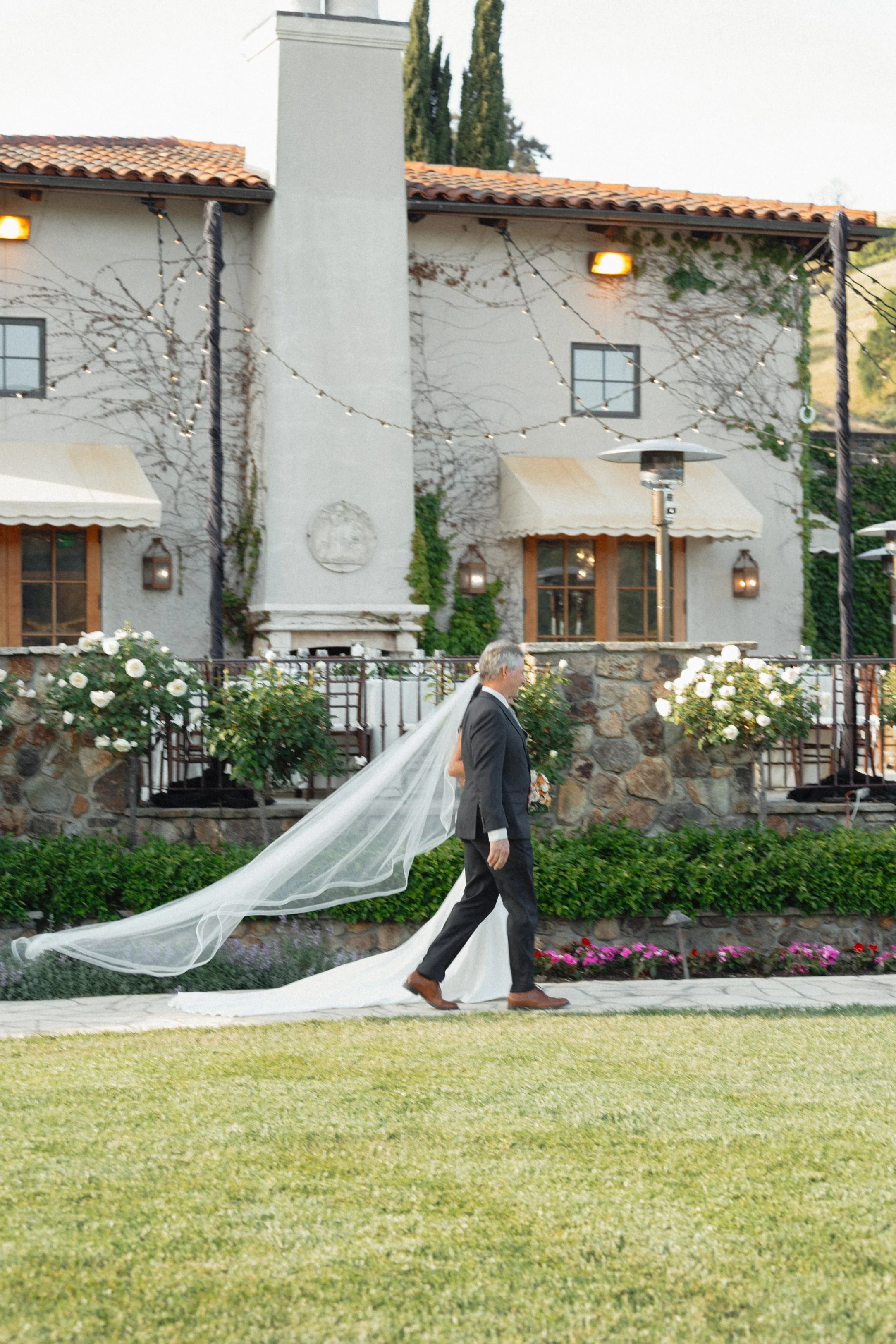 A bride and groom walking outdoors on a lawn in front of a white house with string lights, flowers, and a stone wall.