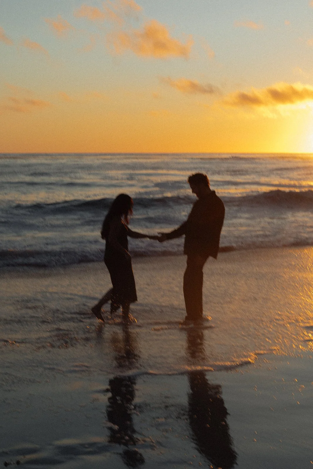 A silhouette of a man and woman holding hands and facing each other while standing in the shallow water on a beach at sunset, with the ocean and clouds in the background and their reflections on the wet sand.