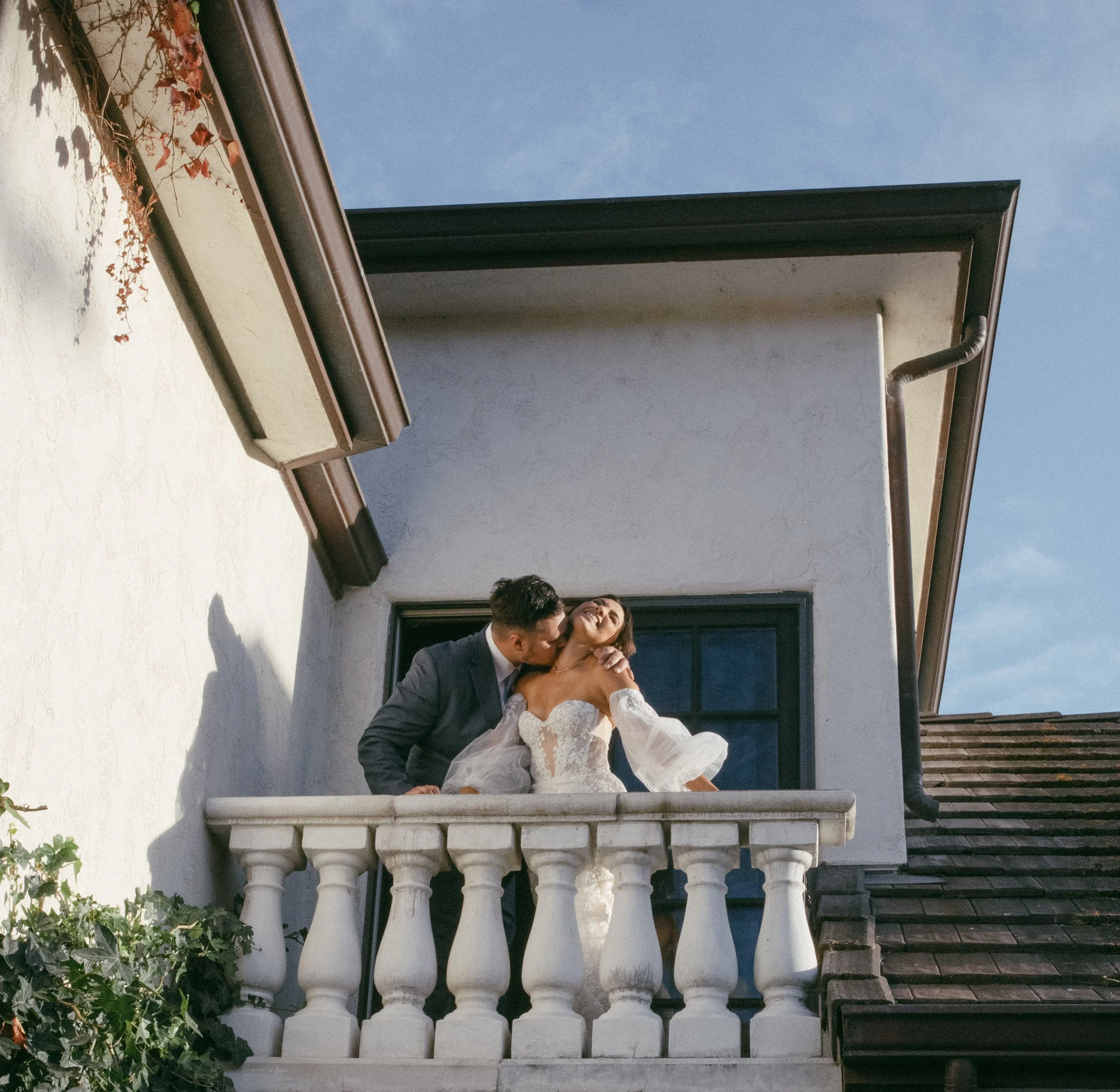 A bride and groom on a balcony. The groom is kissing the bride on the cheek, and she is smiling with her eyes closed. The bride is wearing a white wedding dress, and the groom is in a dark suit. The building has white walls, a window behind them, and