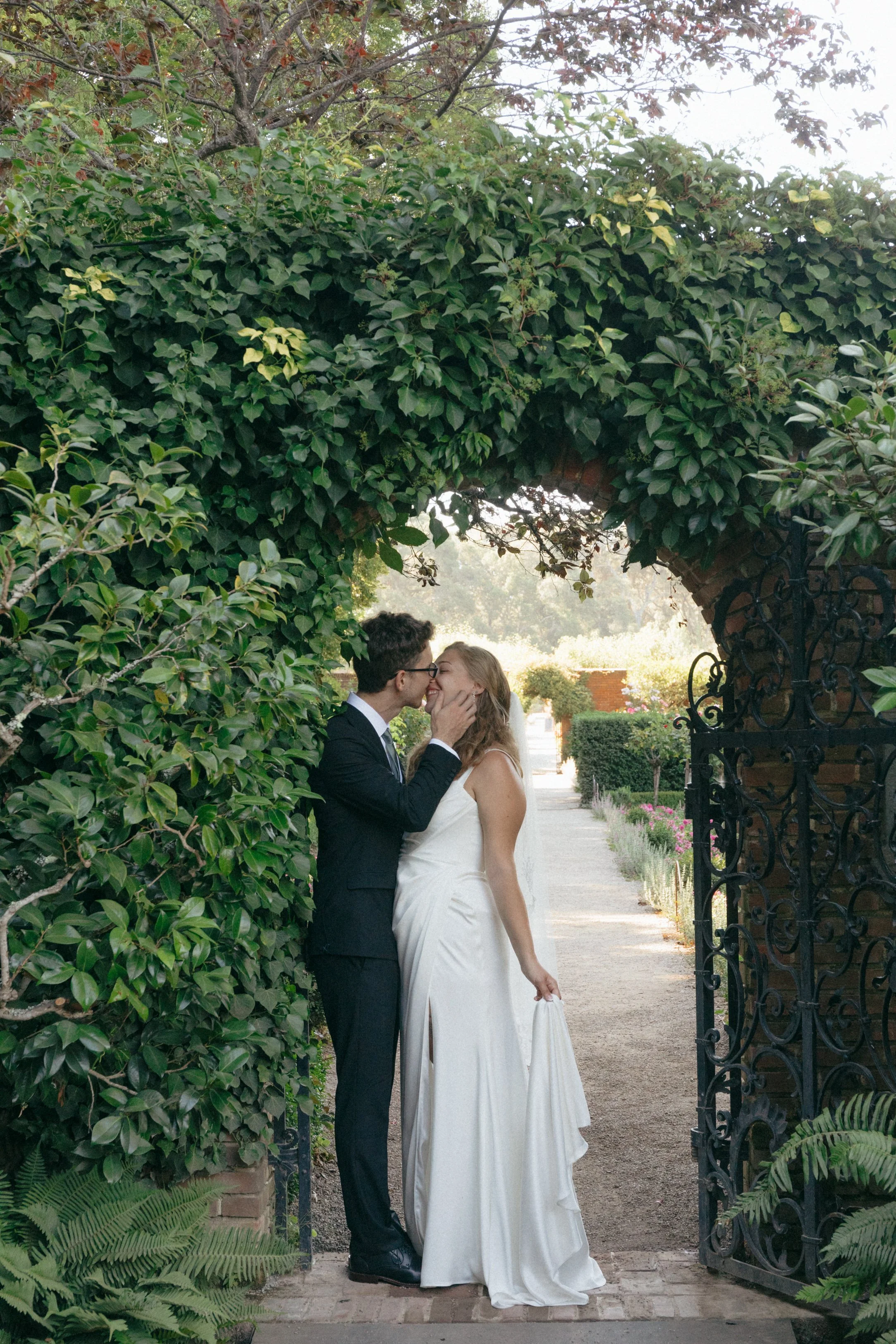 A newlywed couple sharing a kiss in front of a lush, green, leafy archway.