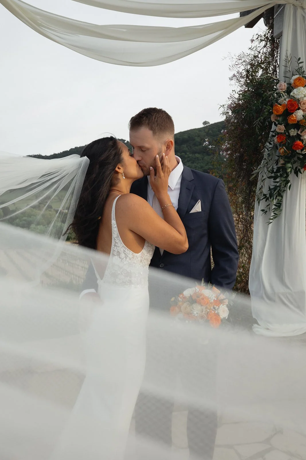 A bride and groom kissing during their outdoor wedding ceremony, with a scenic mountainous background and floral decorations.