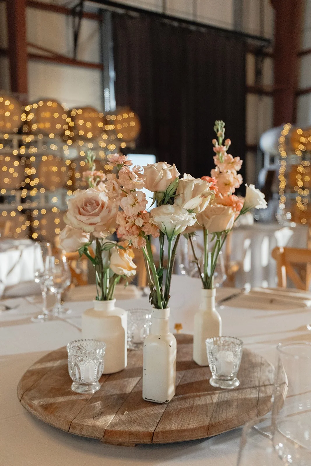 A decorated table with three white vases holding pink and white flowers on a round wooden centerpiece, surrounded by glass candle holders in a warmly lit setting with blurred string lights and black curtains in the background.