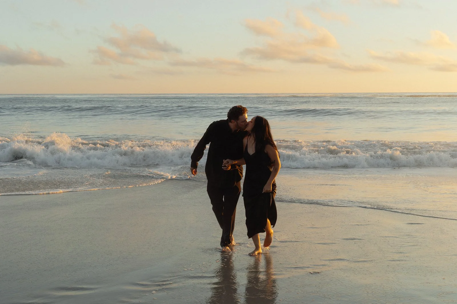 A couple dressed in black walking on the beach at sunset, sharing a kiss.