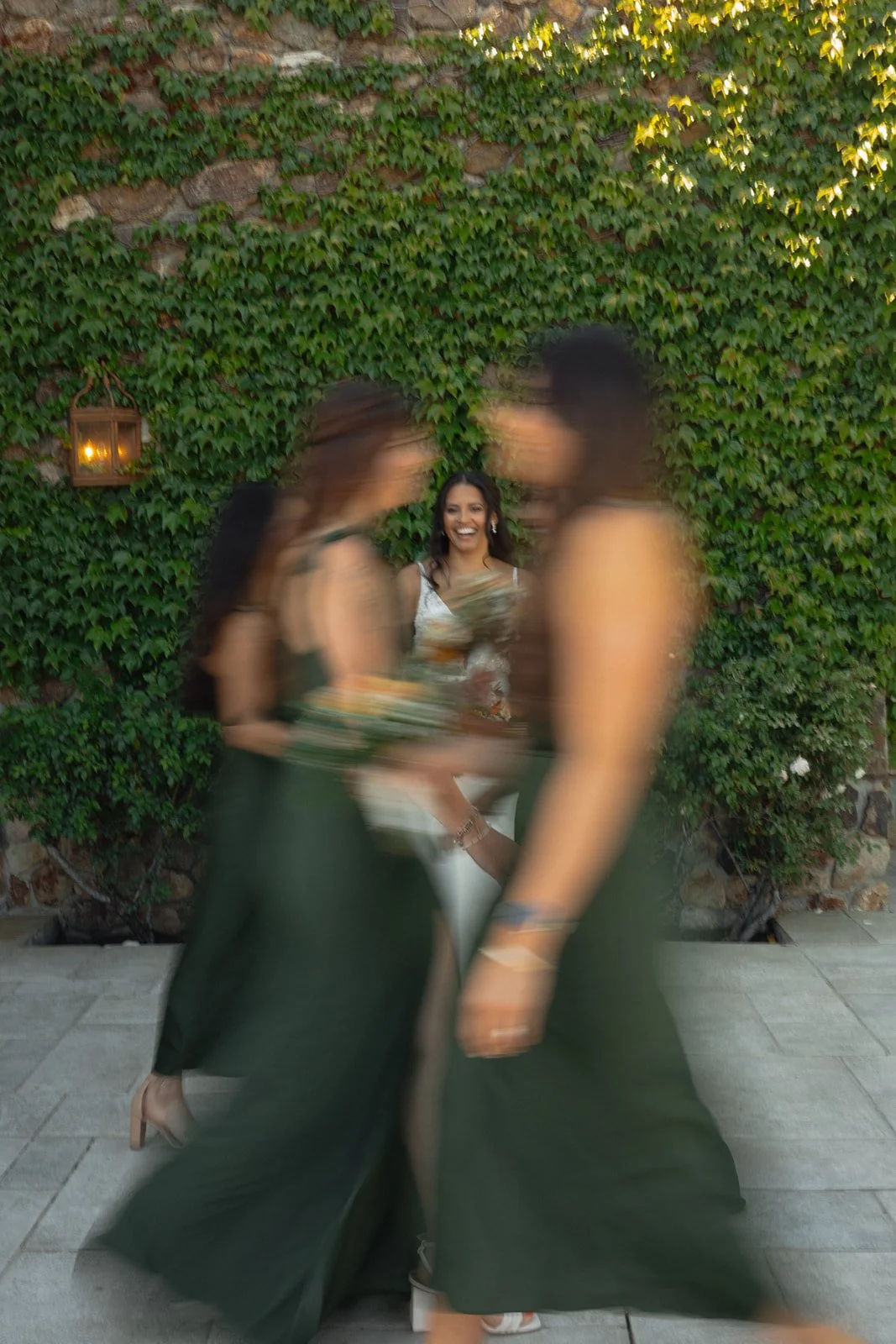 A woman in a white dress smiling and standing in front of a green ivy-covered wall, with two women in dark dresses walking past in a blurred motion.