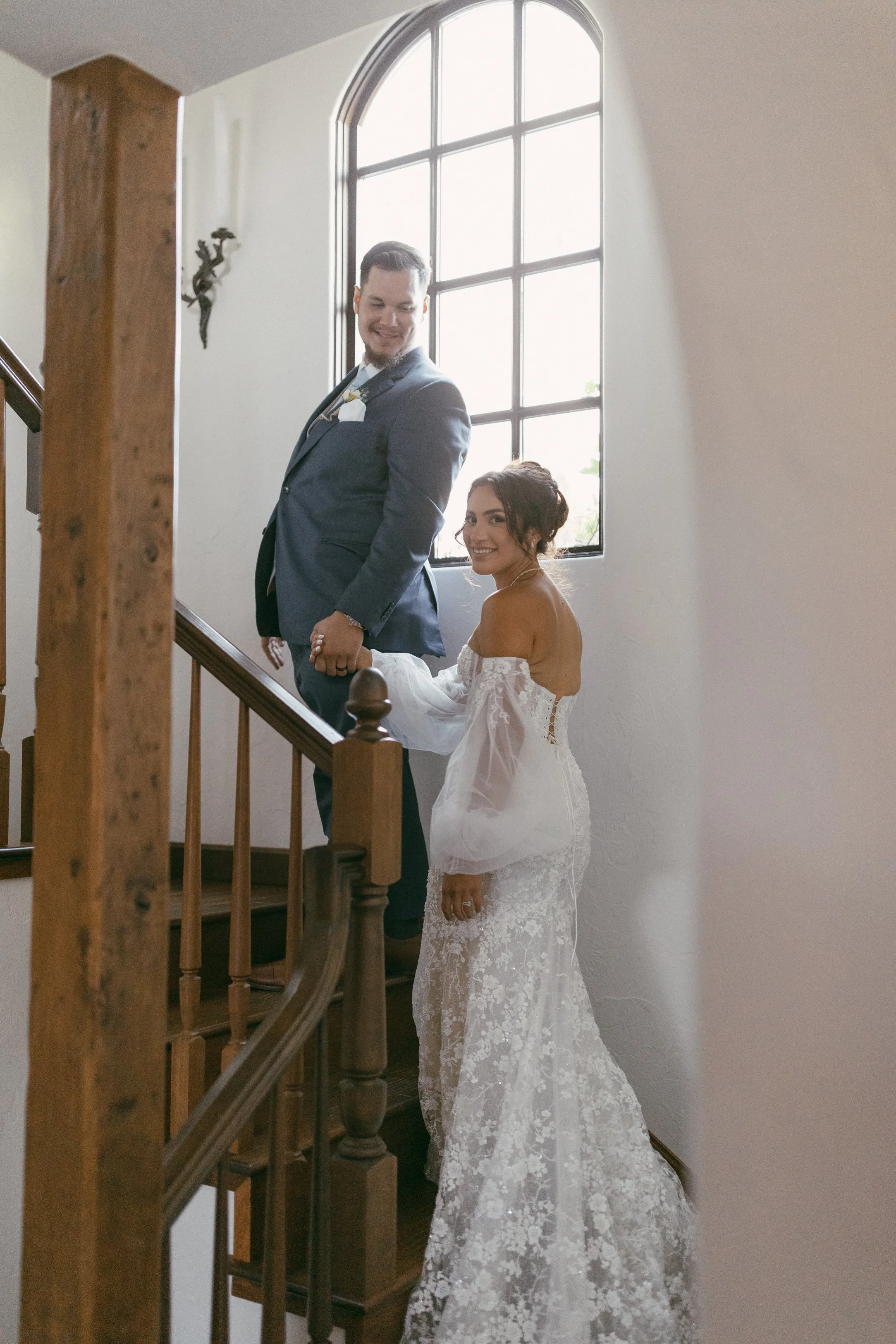 Bride and groom standing on staircase near large window, smiling, with bride in a lace wedding gown.