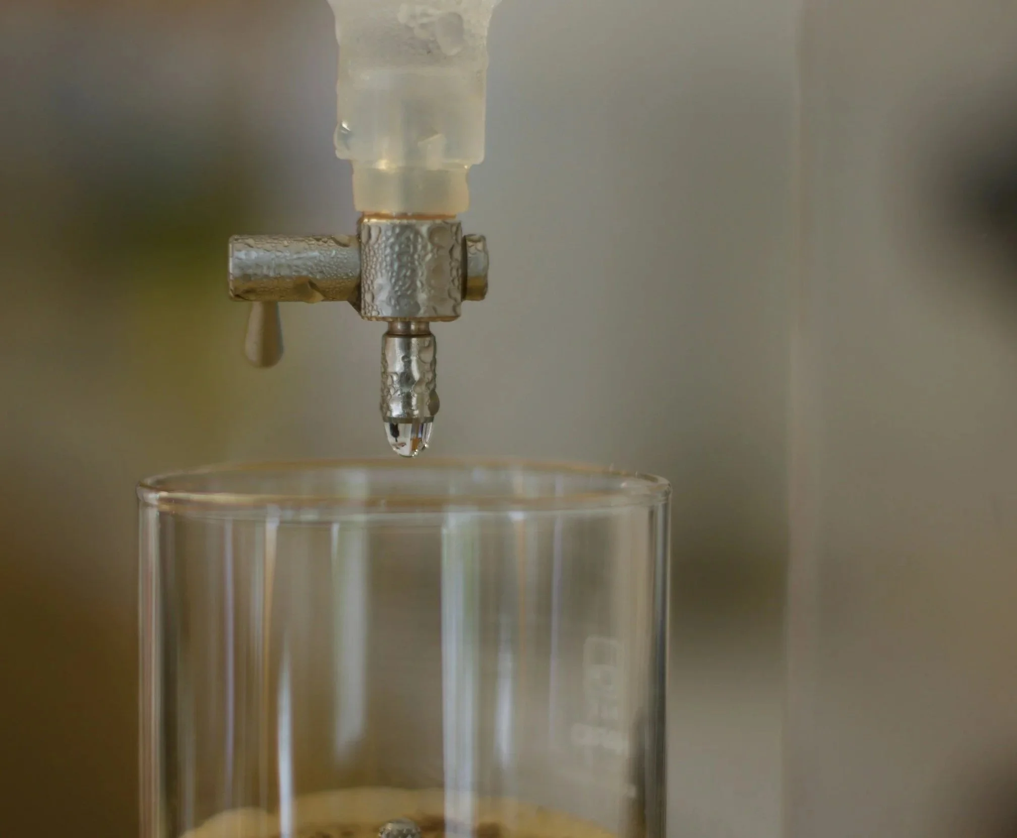 Close-up of a water droplet falling from a metallic nozzle into a clear glass.