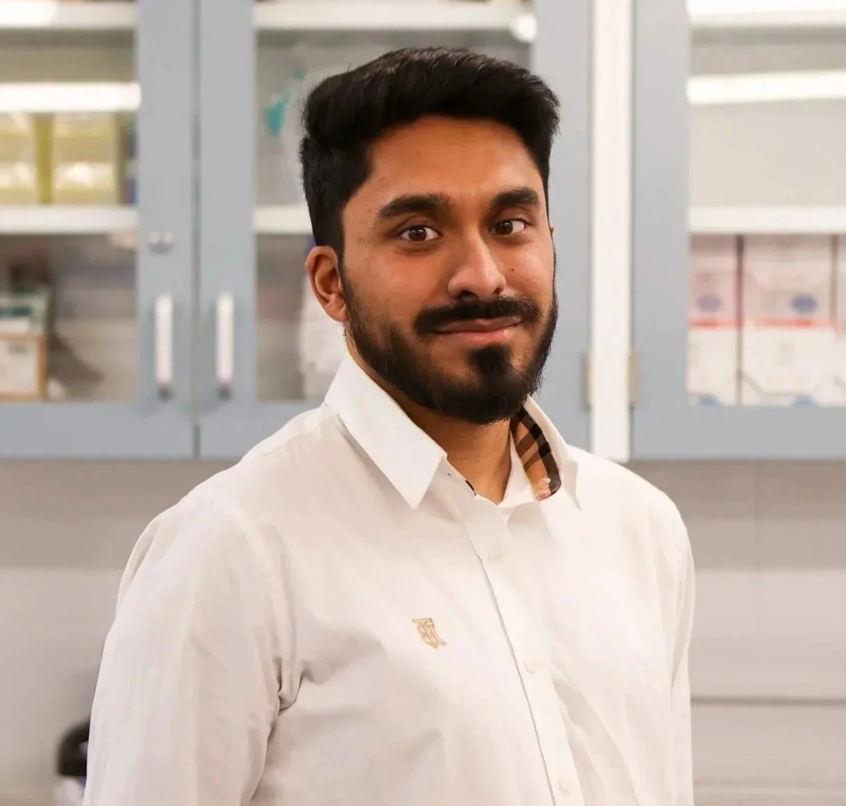 A man with dark hair and a beard, smiling, wearing a white shirt, standing outdoors with pink flowers and green foliage in the background.