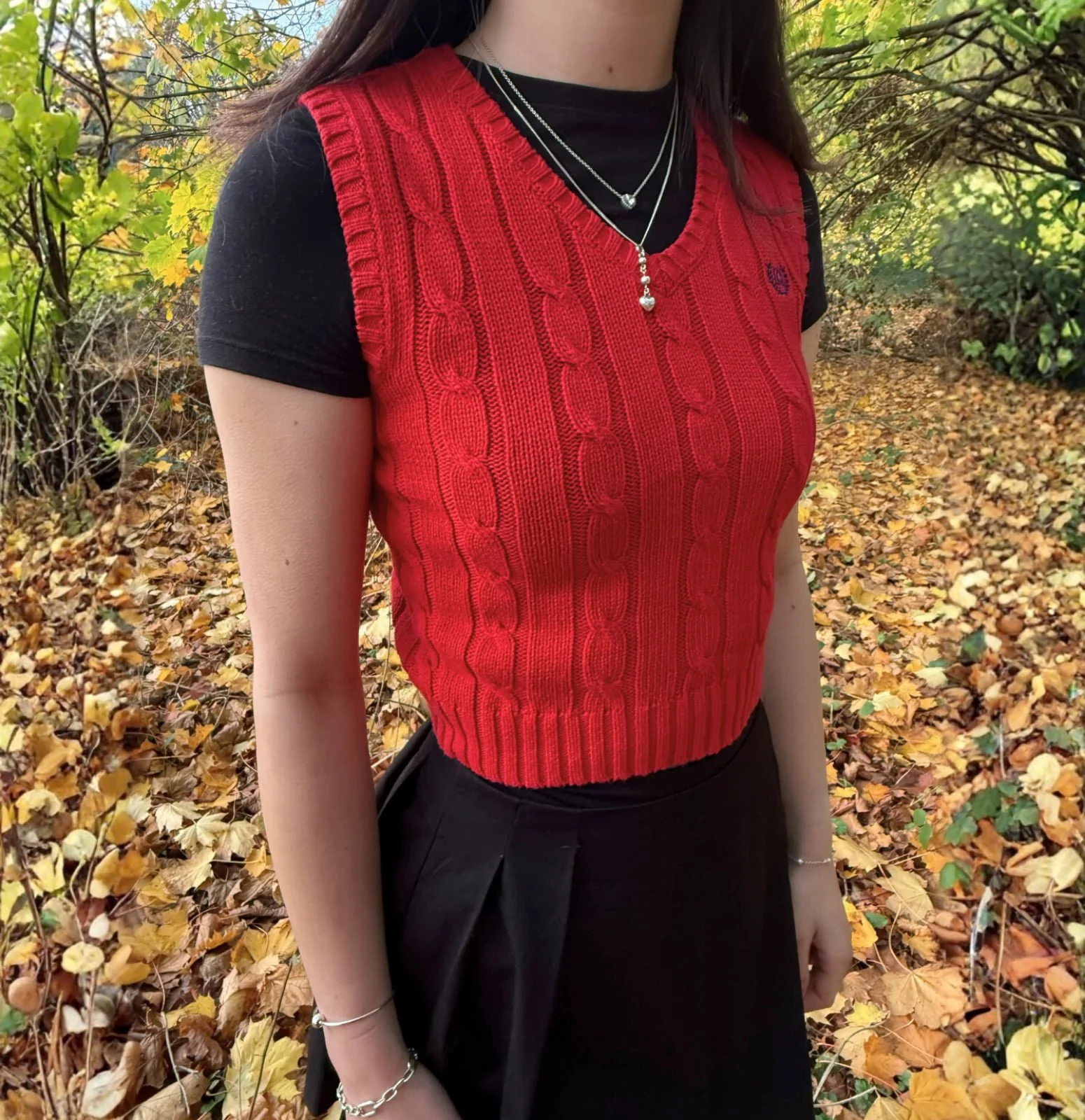 A woman wearing a red cable knit vest over a black t-shirt stands outdoors in an autumn setting with yellow and brown leaves on the ground and trees with yellowing foliage in the background.