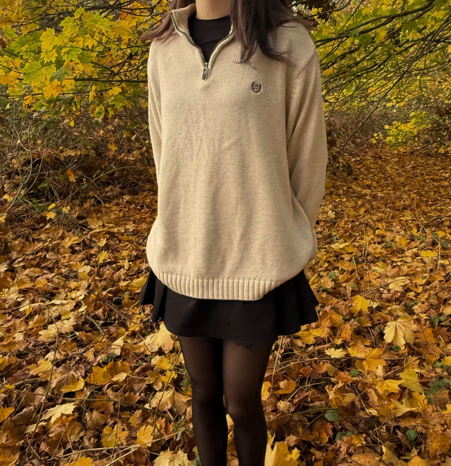 Person standing outdoors on a bed of fallen autumn leaves, wearing a light beige quarter-zip pullover, black skirt, and black tights.