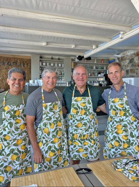 Fyra män i kök, alla iklädda aprikosmönstrade förkläden med citronmönster, står bredvid varandra på en matlagningskurs i Sicilien.