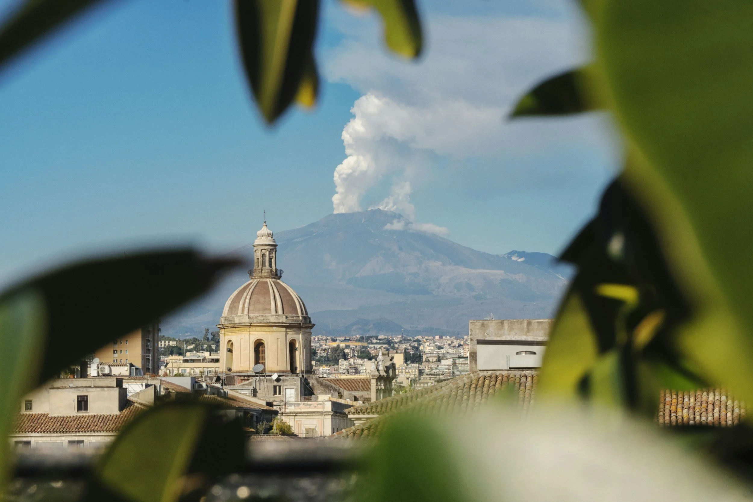Utsikt över en stad med en kupolformad byggnad i förgrunden, ett berg med en rykande vulkan i bakgrunden och löv runt kanterna. Vulkanen Etna strålar vackert över östra Sicilien.