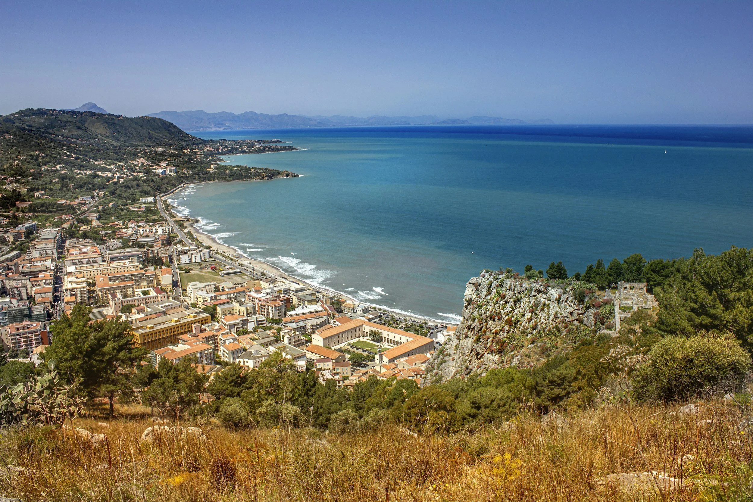 En kuststad med en strand under en solig dag, omgiven av gröna kullar och berg i fjärran i Macari, Sicilien.