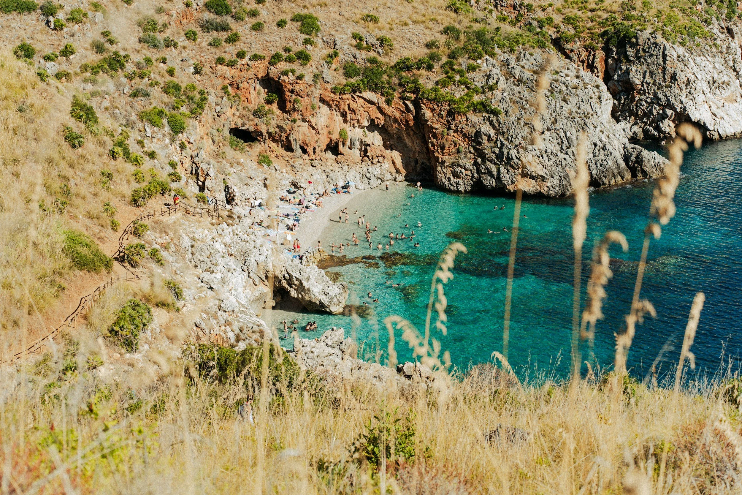 A secluded beach with clear turquoise water, surrounded by rocky cliffs and green shrubbery, with many people swimming and sunbathing.