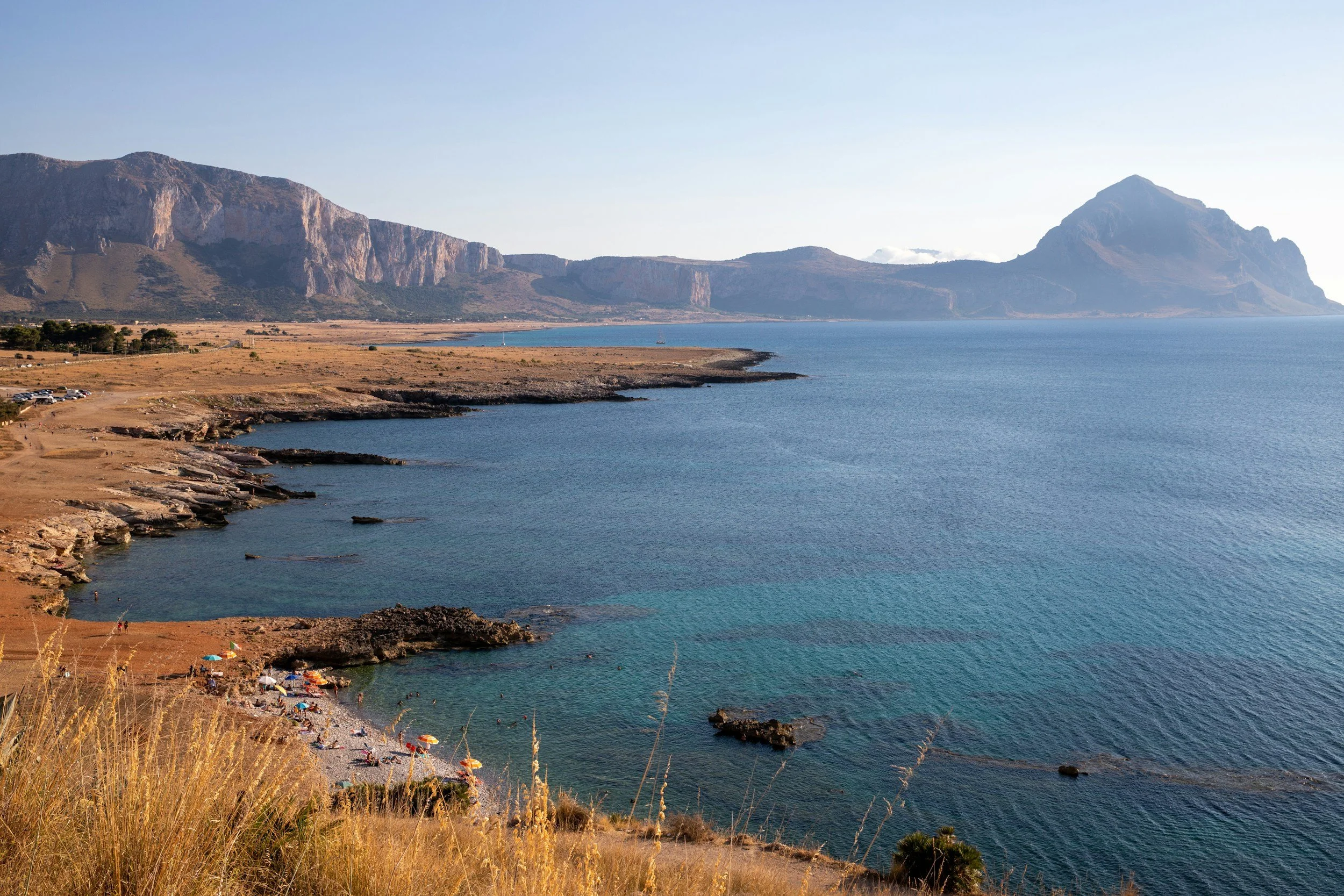 En kustlinje med klippor och en liten strand med solbadare och parasoller, med berg i bakgrunden under en klar himmel i Trapani, Sicilien.