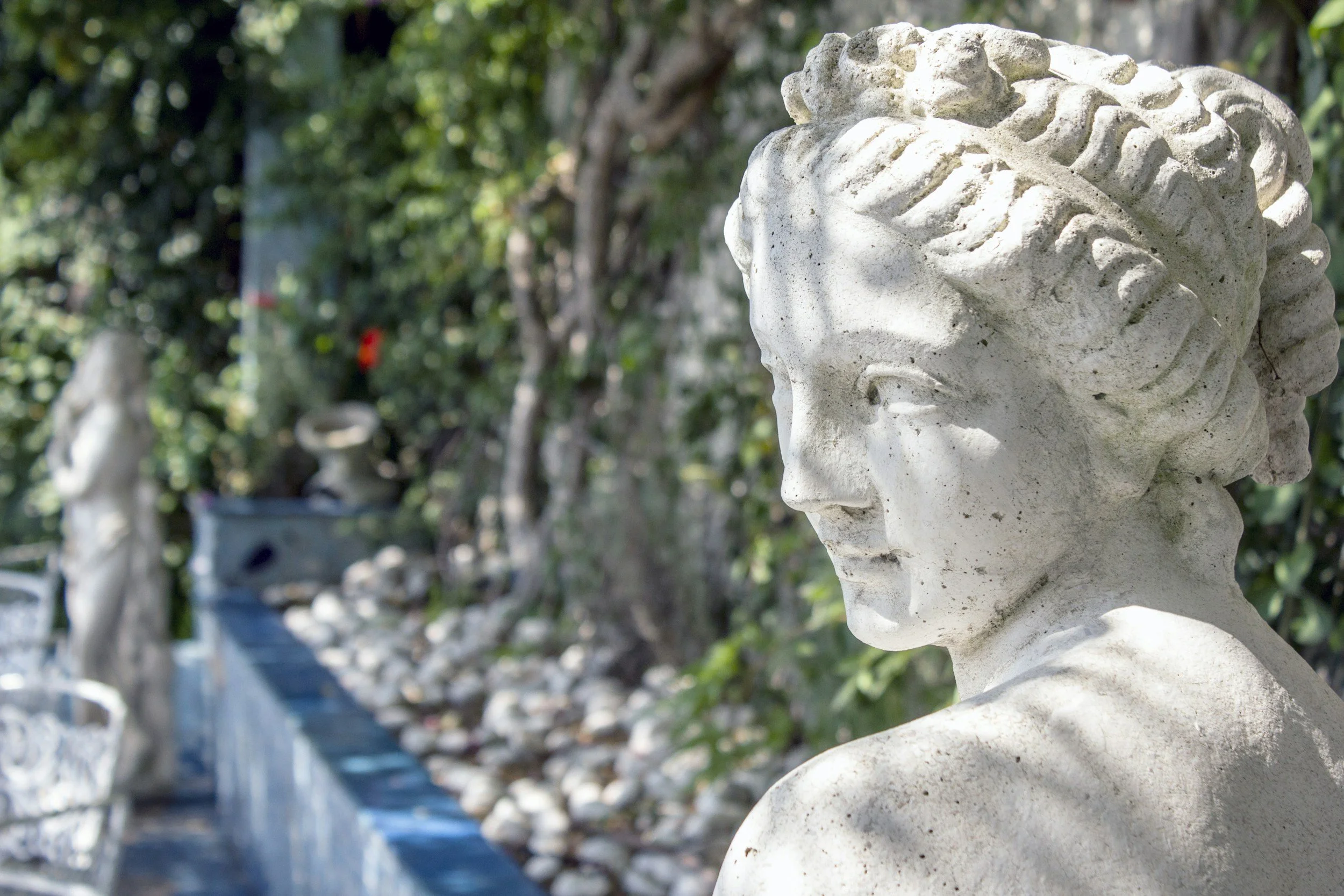 En vit stenen skulptur av en kvinnlig figur med armarna bakom ryggen, placerad utomhus bland gröna växter i Taormina, Sicilien.