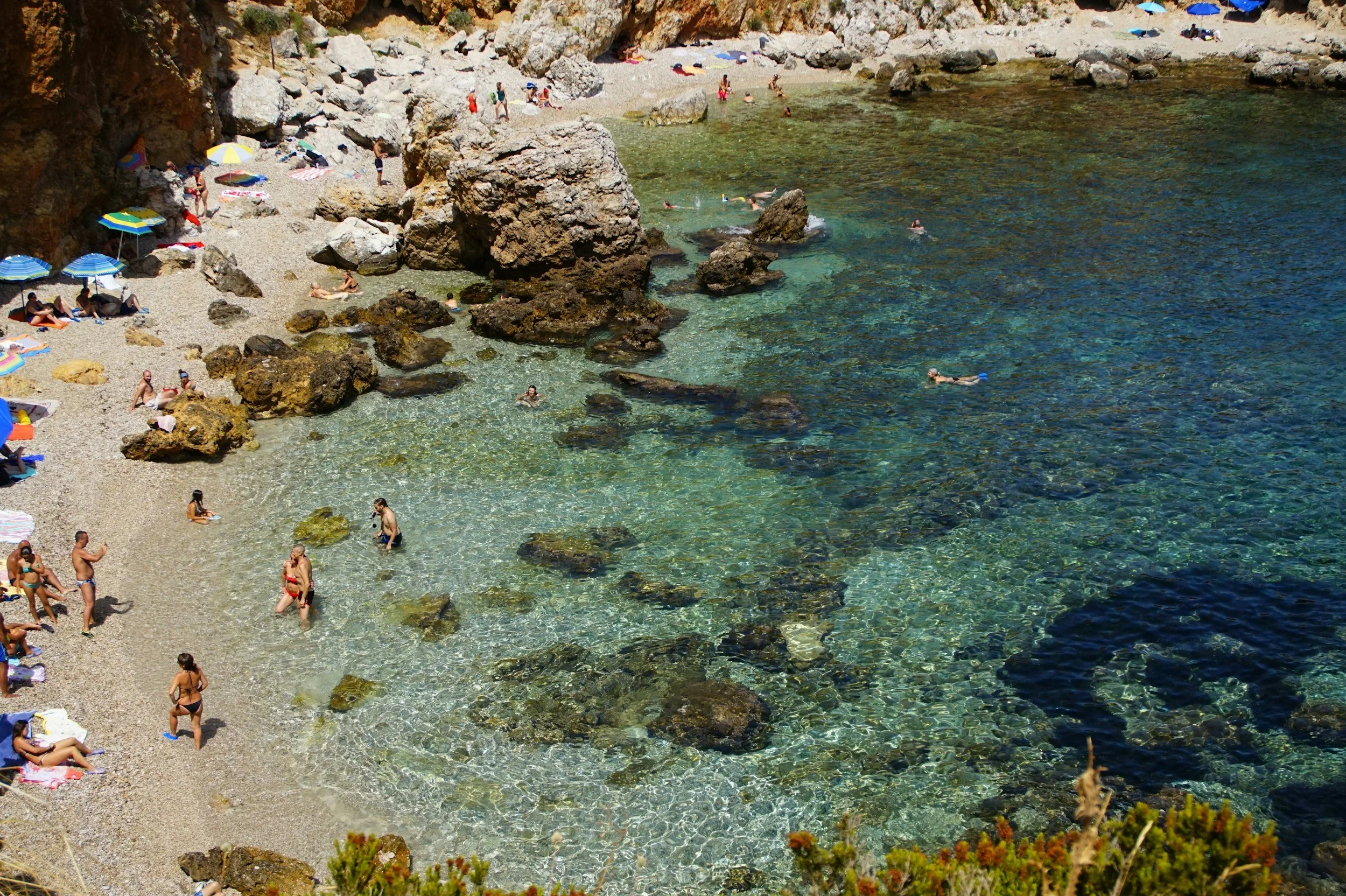 Kustscen i provinsen Trapani, Sicilien med färgglada byggnader, klippiga bergväggar och turkost vatten, med båtar, simmare och solbadare på stranden.