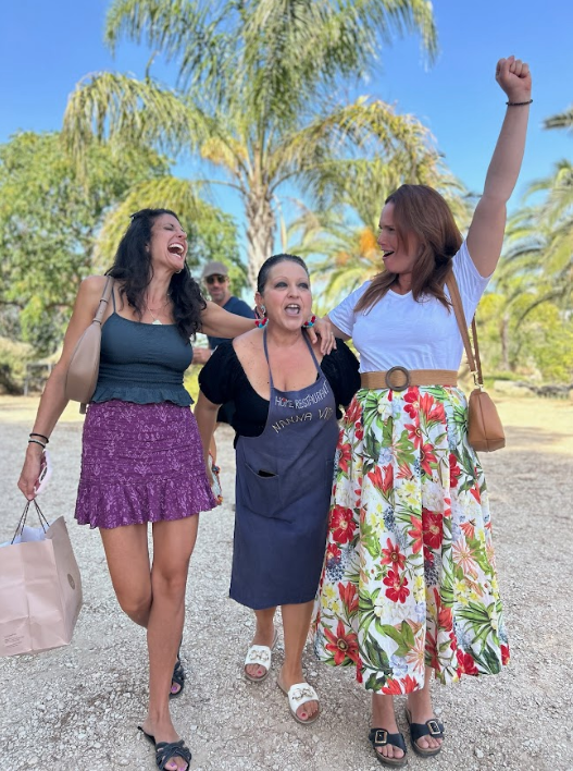 Tre kvinnor som ler och skrattar, två av dem har armarnas om varandra och den tredje har armen i luften, utomhus med palmträd och blå himmel i bakgrunden i Sicilien.