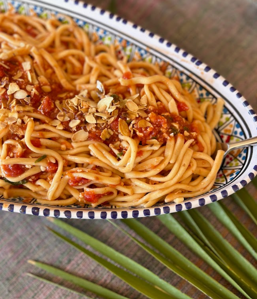 En skål med spagetti i tomatsås toppat med hackade mandlar, placerad på ett bord med gröna blad under skålen i Sicilien.
