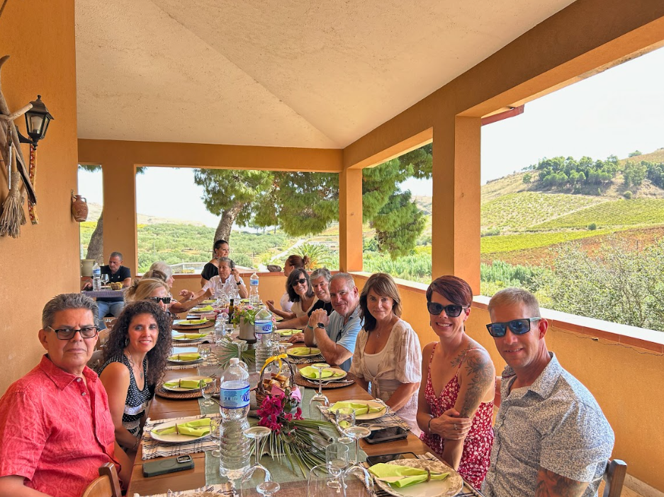 Flera personer som sitter vid ett bord på en veranda med utsikt över vinrankor och kullar i Sicilien.