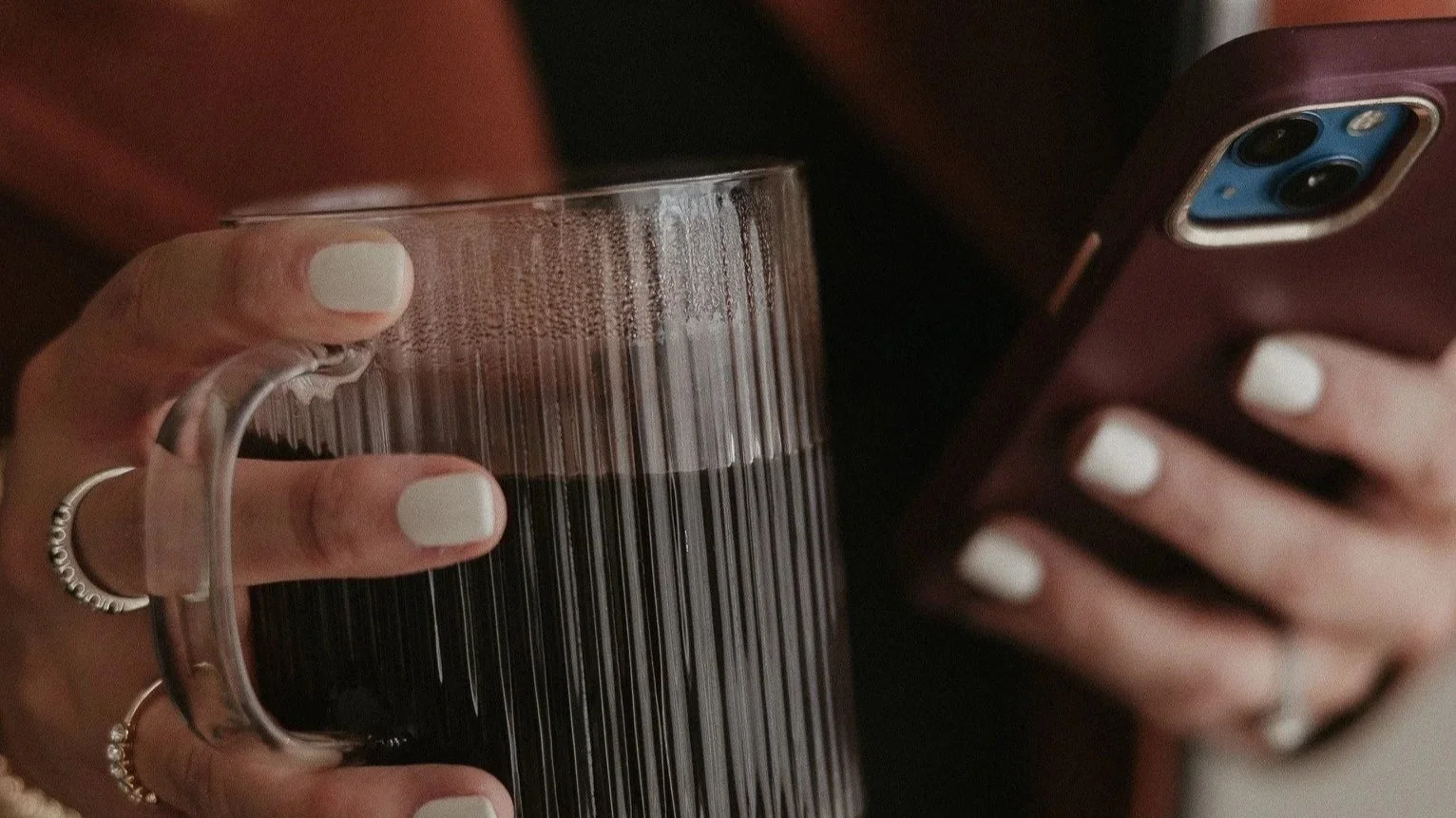 Close-up of a person holding a glass mug filled with a dark beverage, and a smartphone with a purple case in the other hand. The person has white painted nails and is wearing rings.