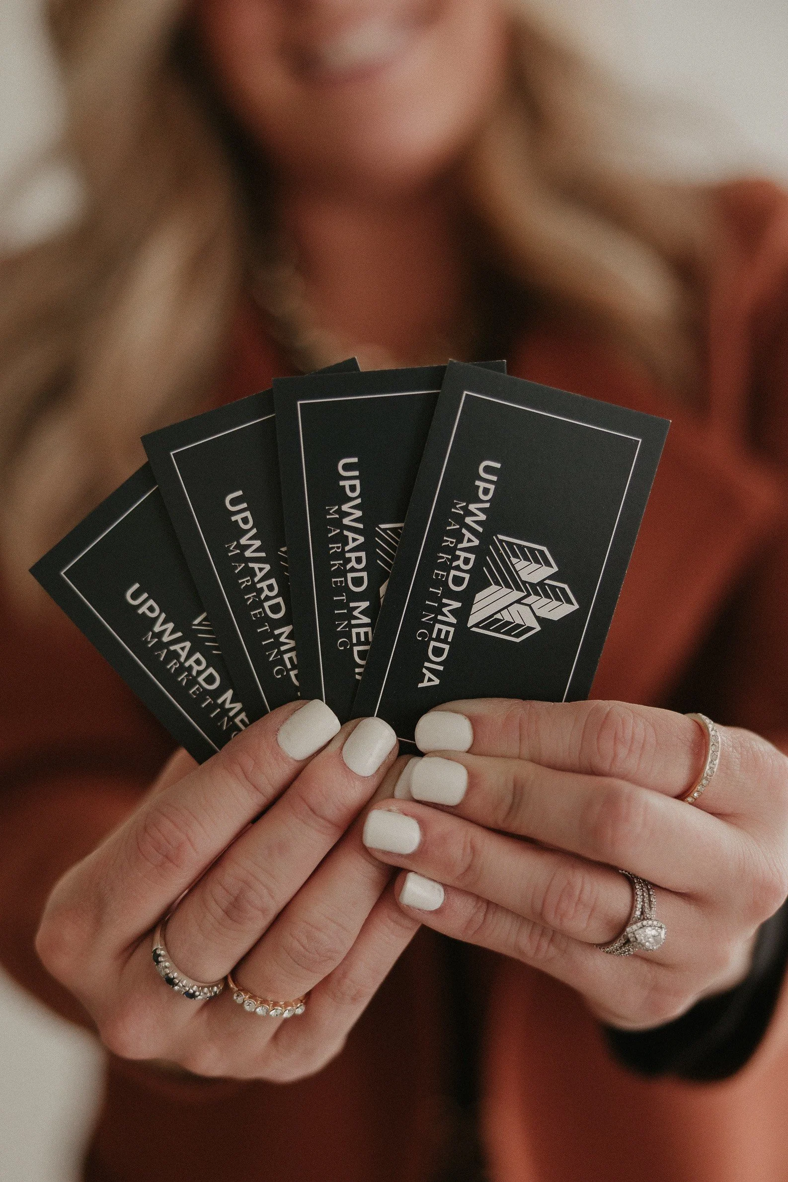 A woman with blonde hair holding five Upward Media marketing business cards with white and silver rings on her fingers and in front of a blurred background.