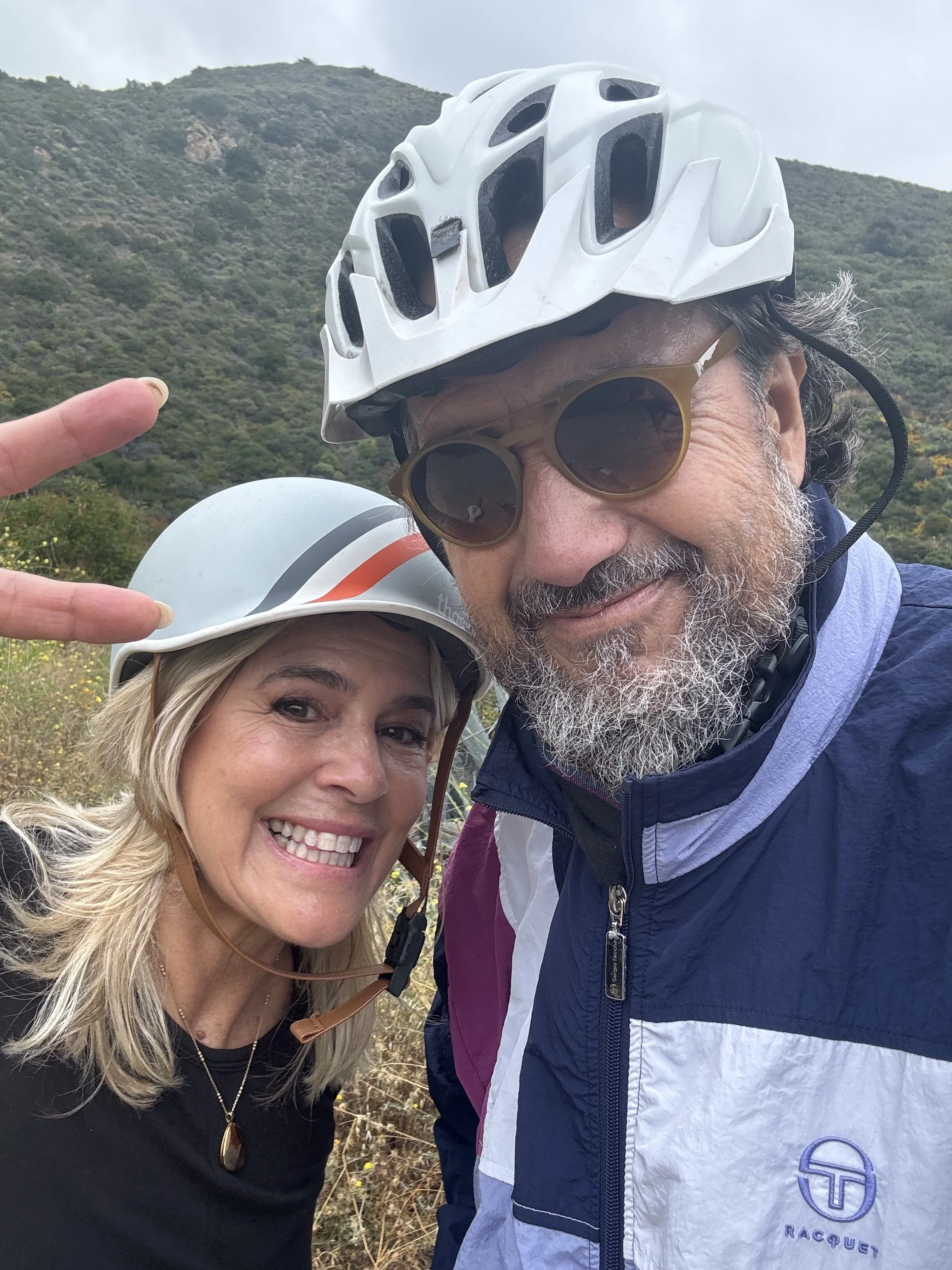 Two people in cycling gear wearing helmets, smiling outdoors with a hilly, green landscape in the background.