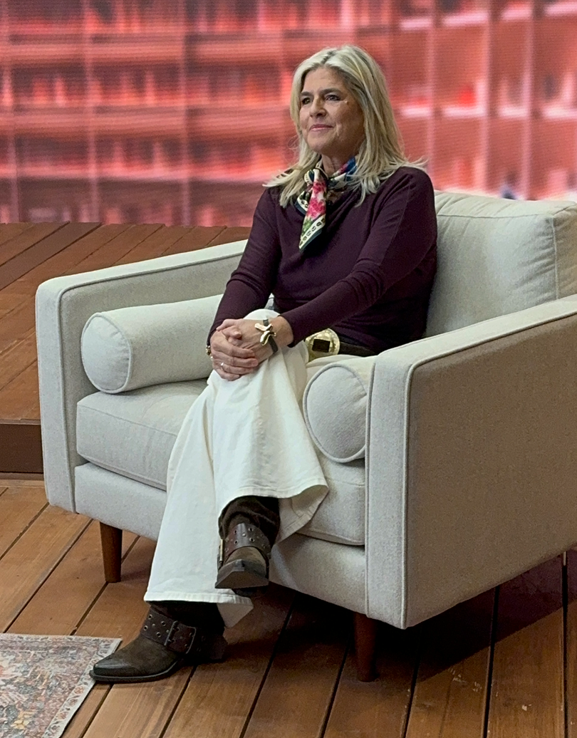A woman sitting on a white armchair with a beige cushion, wearing a dark long-sleeve shirt, white wide-leg pants, and brown boots, with a patterned scarf around her neck, on a wooden floor with a red background.