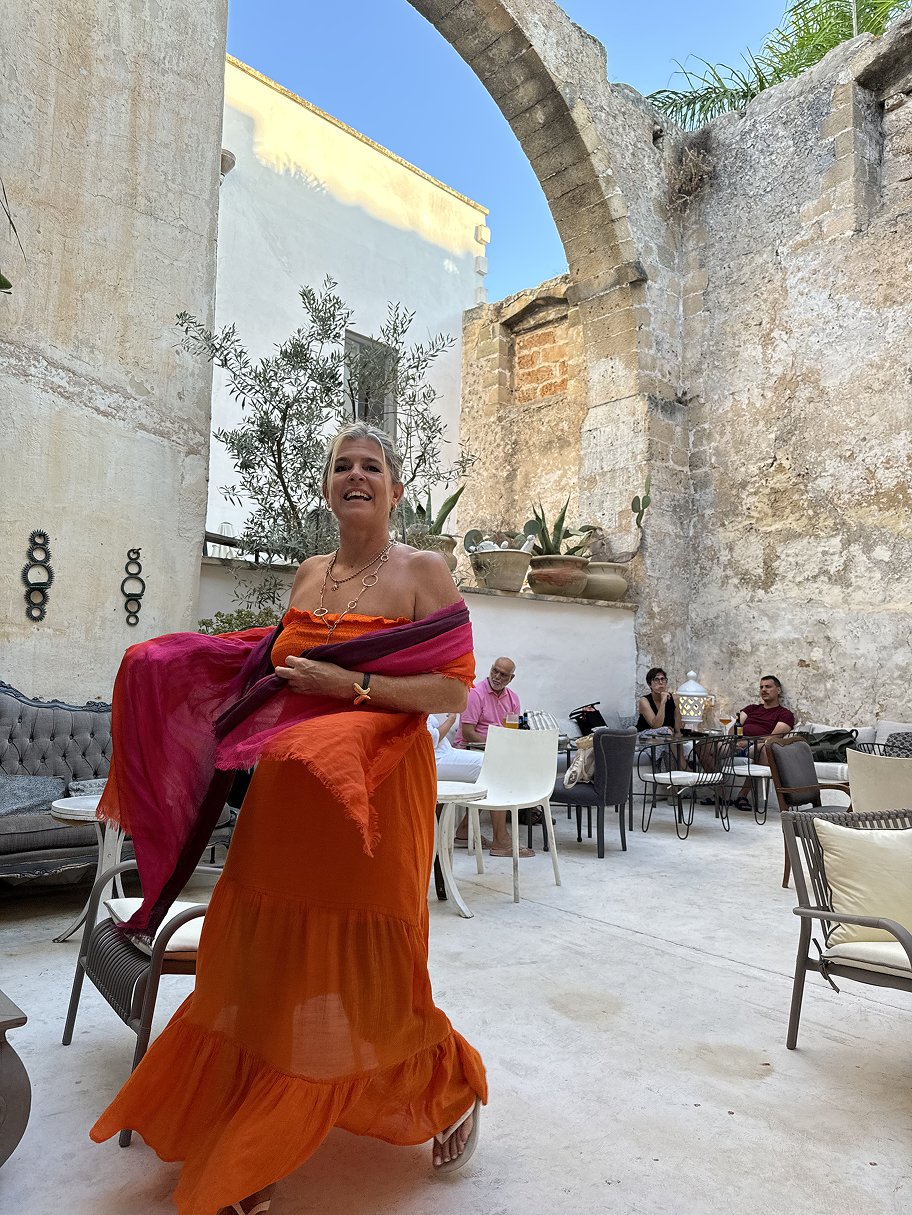 A woman in an orange dress smiling and holding a pink and purple shawl, standing in a courtyard with a few people sitting at tables behind her, surrounded by old stone walls and potted plants.
