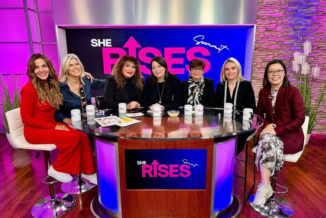 Seven women sitting around a TV studio table with mugs, a bowl, and papers, celebrating the show 'She Rises.'