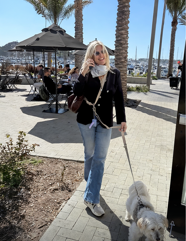 A woman with blonde hair, sunglasses, and a white scarf, smiling while talking on her phone, walking her small white dog on a leash near a marina with boats, palm trees, and outdoor seating in sunny weather.