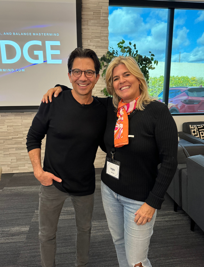 A man and woman standing together inside a modern office space, smiling at the camera. The man has dark hair, glasses, and is wearing a black long-sleeve shirt and gray pants. The woman has blonde hair, a colorful scarf, and is dressed in a black sweater and light jeans with a ripped knee. Behind them is a large window with a view of a parking lot and greenery, and a presentation screen displaying a logo or presentation slide.
