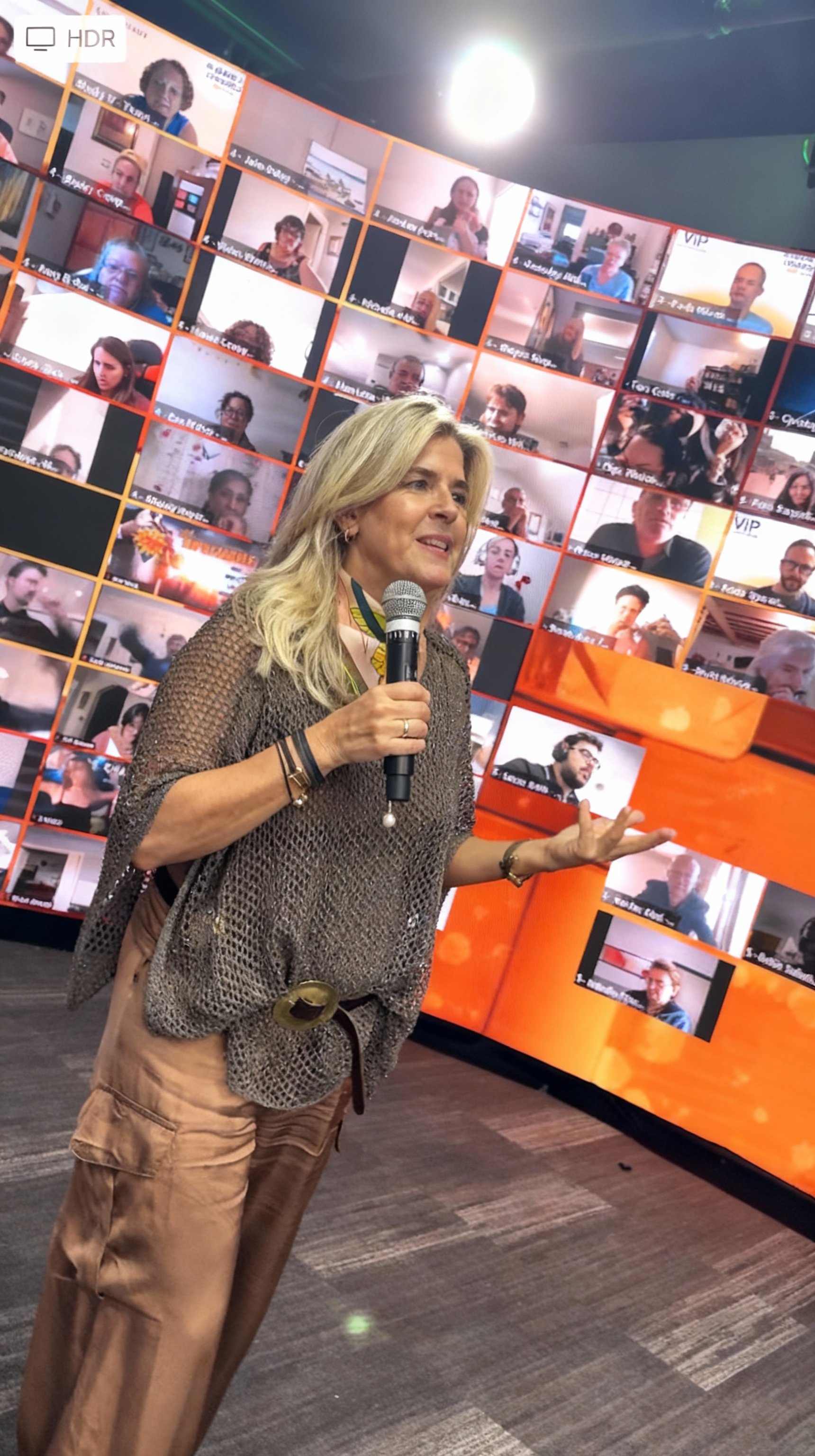 A woman with blonde hair holds a microphone and speaks in front of a large digital screen displaying a grid of virtual participants in a video conference.