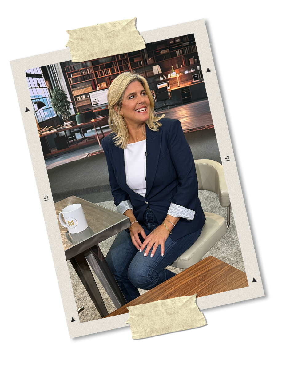 A woman with blonde hair wearing a navy blazer and white shirt smiling and sitting at a table with a white mug in a modern office setting.
