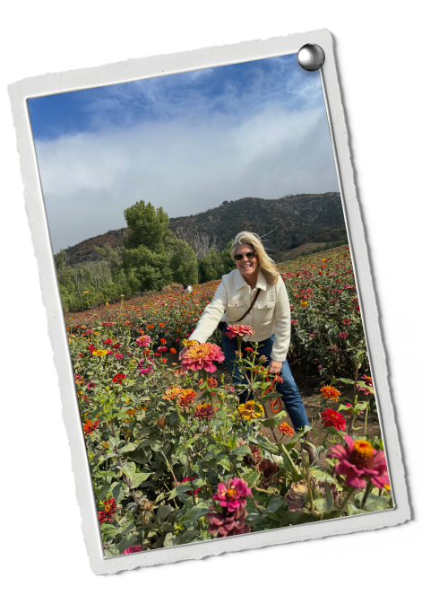 A woman with blonde hair wearing sunglasses, a white jacket, and blue jeans, is standing in a colorful flower field with red, pink, and orange flowers, with green hills and mountains in the background under a partly cloudy sky.