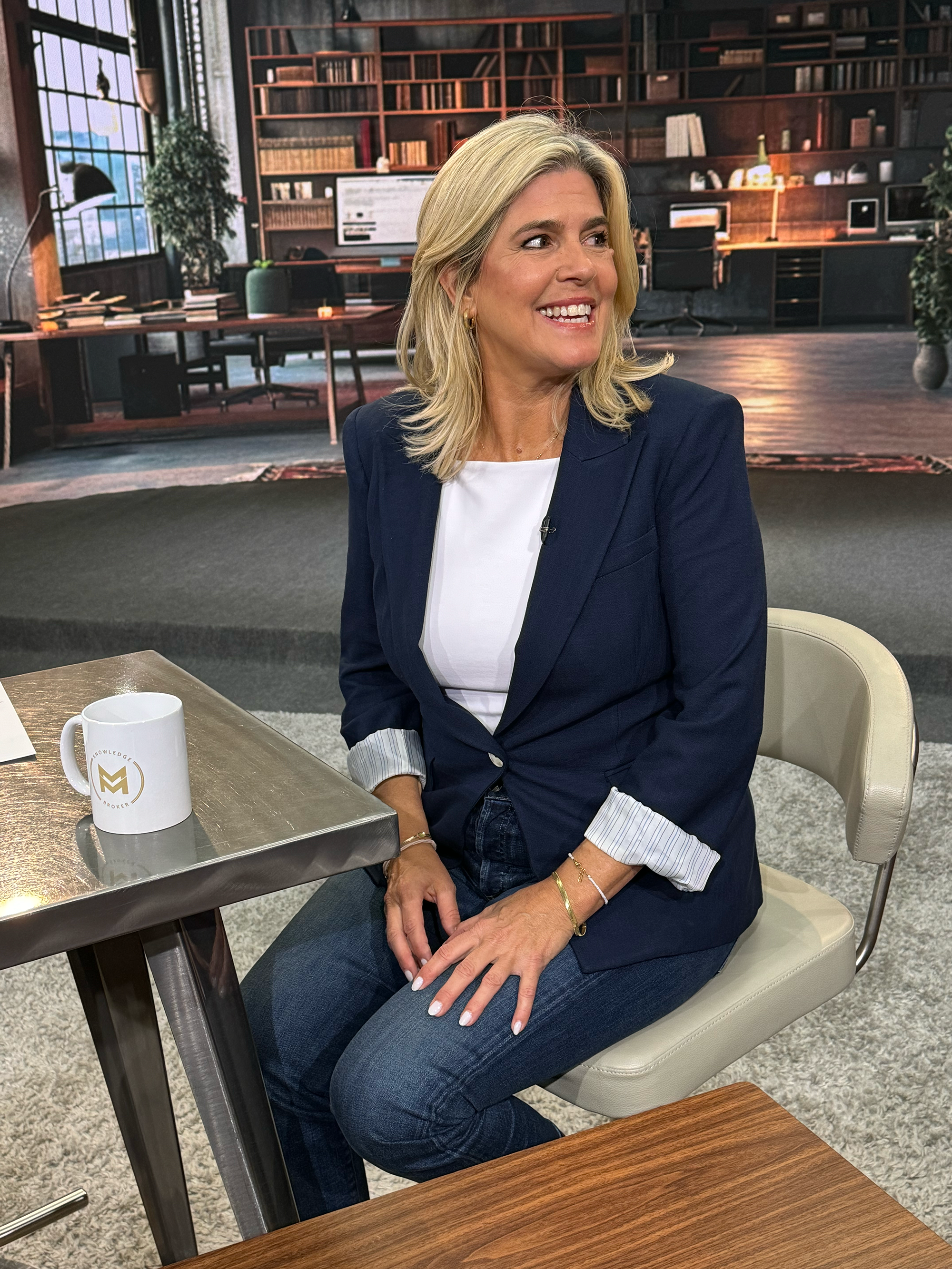 A woman with blonde hair sitting in a cream-colored chair at a desk in a studio, with a large bookshelf and office setting in the background, wearing a navy blazer over a white top, smiling and looking to her right.