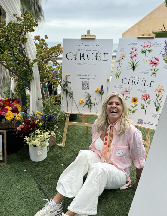 Woman sitting on grass at flower garden event, smiling, surrounded by flowers and botanical posters.