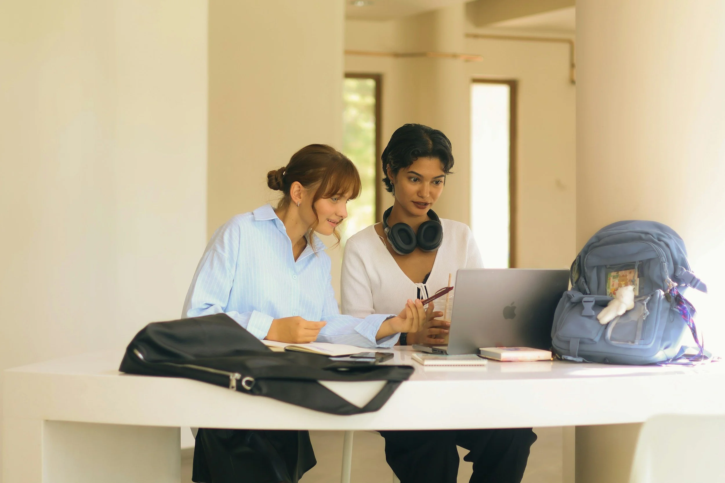 Two women sitting at a white table, looking at a laptop, with backpacks and notebooks. One woman has headphones around her neck, and they are in a bright room with large windows.